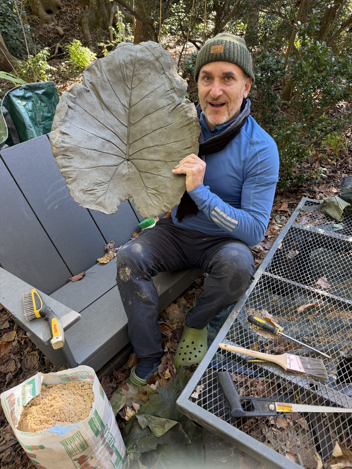 The maker sitting outdoors holding up a freshly freed concrete leaf cast, tools all around